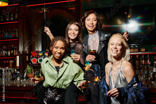 cheerful stylish multiracial girlfriends with cocktails and shiny sparklers looking at camera in bar
