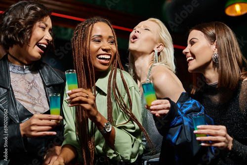 overjoyed and cheerful multicultural girlfriends holding shot glasses in bar, festive party