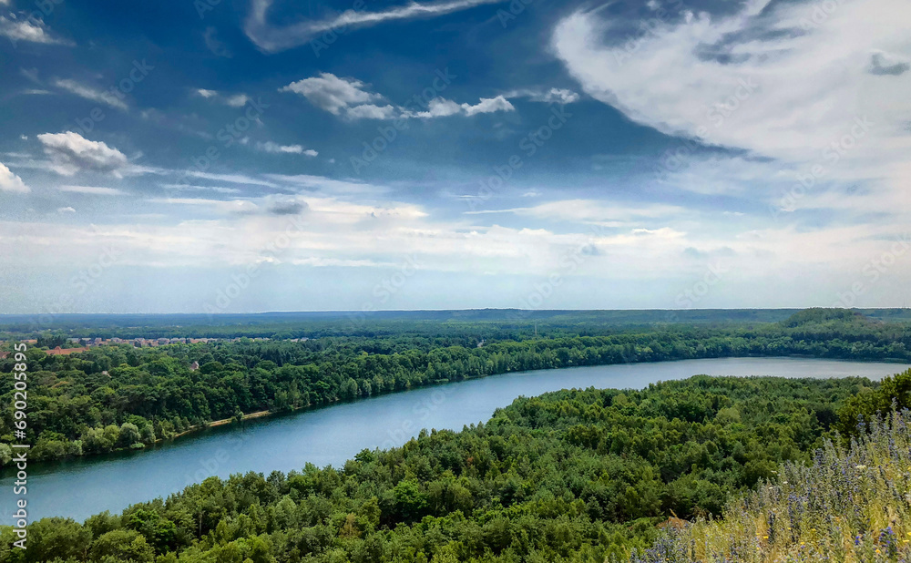 This landscape photograph offers a breathtaking panoramic view of a meandering river cutting through a lush green forest. The expansive blue sky, adorned with wispy clouds, crowns the scene