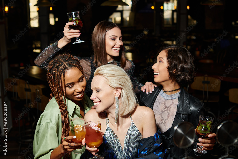 cheerful elegant women clinking cocktail glasses near multiethnic girlfriends during party in bar