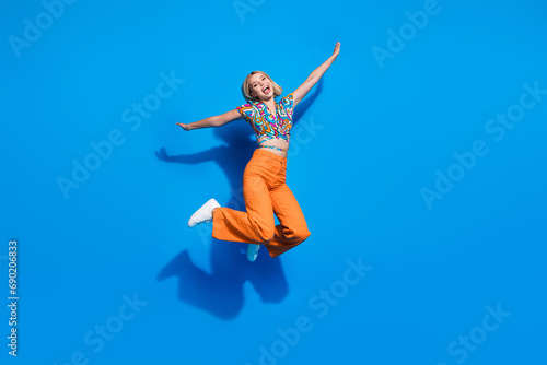 Full size photo of cute good mood girl wear blouse orange trousers jumping holding palms like wings isolated on blue color background