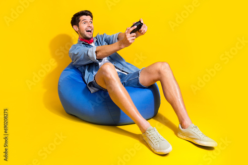Portrait of impressed man dressed denim shirt sit on bean bag hold joystick look at game empty space isolated on yellow color background