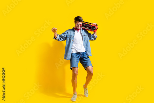 Full body photo of positive man dressed denim shirt shorts in glasses dance with boombox enjoy music isolated on yellow color background