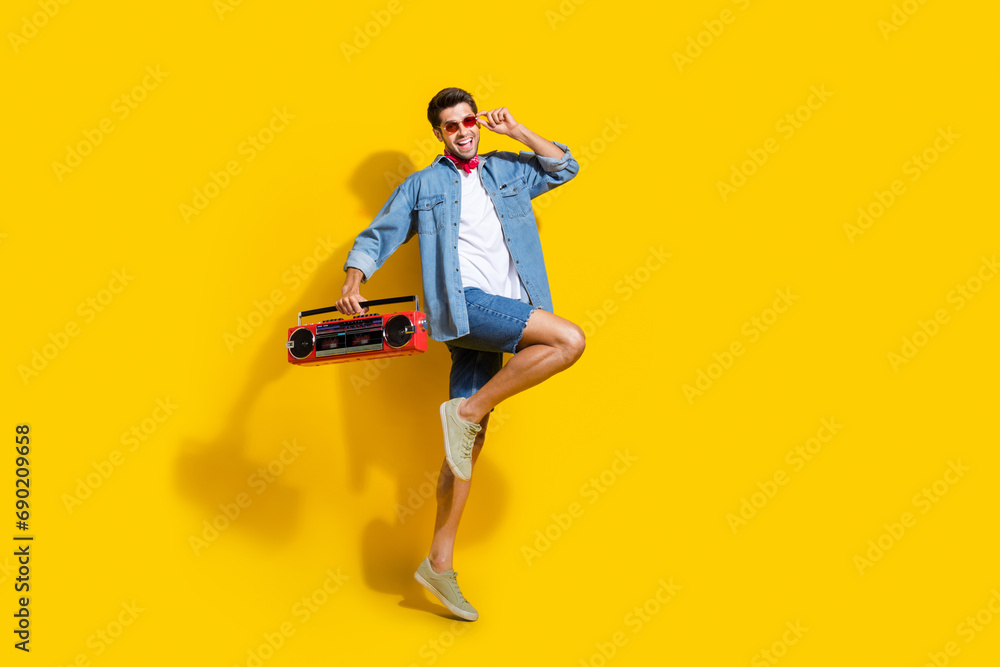 Full body photo of cool man dressed denim shirt flying touching sunglass hold boombox at party isolated on yellow color background Full body photo of cool man dressed denim shirt flying touching sunglass hold boombox at party isolated on yellow color background