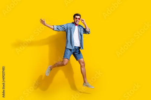 Full body photo of satisfied pleasant man dressed denim shirt shorts flying touching sunglass isolated on bright yellow color background