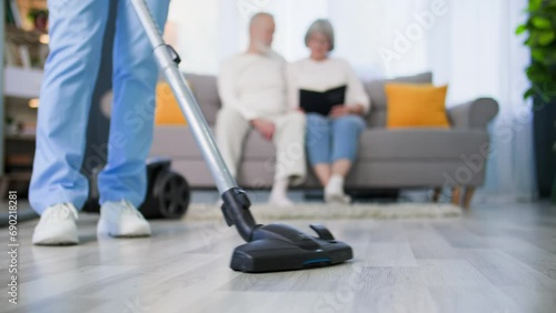 caring about elderly people, social worker in a medical uniform with a vacuum cleaner in her hands helps an elderly couple with cleaning house, close-up photo