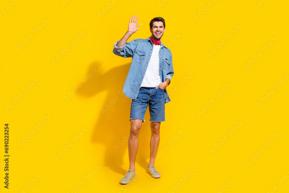 Full body photo of cheerful nice man dressed denim shirt waving palm say hello hand in pocket isolated on vivid yellow color background Full body photo of cheerful nice man dressed denim shirt waving palm say hello hand in pocket isolated on vivid yellow color background