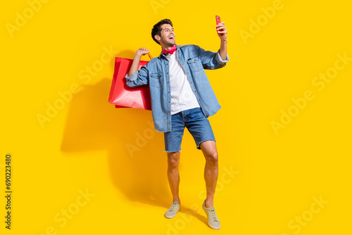 Full body photo of cool man dressed denim shirt shorts holding new outfit in bags look at smartphone isolated on yellow color background.