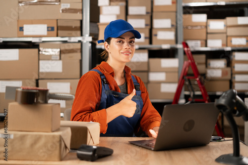 Manager checking online orders and inventory on a laptop while sitting at a desk working in warehouse with shelves full of parcels, showing thumb-up
