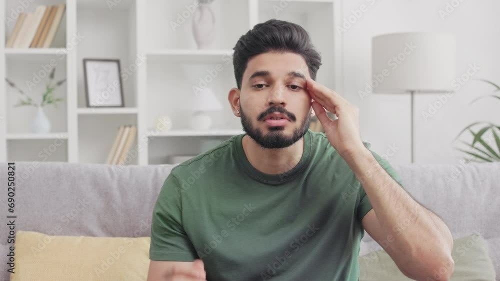 Casually dressed exhausted man with beard touching forehead while sitting on couch and having migraine. Indian young guy feeling tired and stressed staying alone at home. Casually dressed exhausted man with beard touching forehead while sitting on couch and having migraine. Indian young guy feeling tired and stressed staying alone at home.