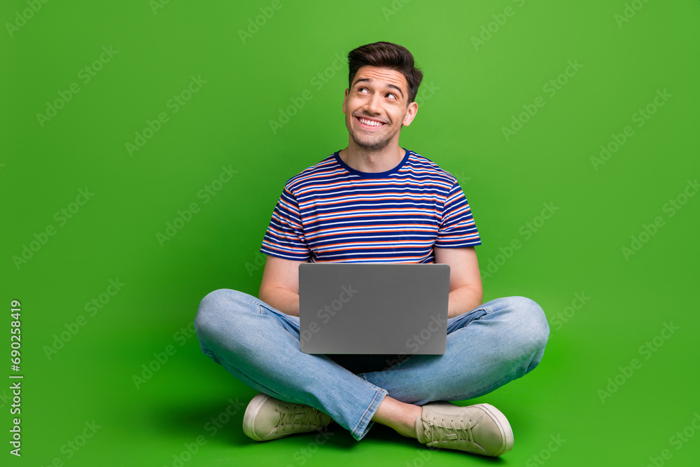 Full size photo of pleasant guy dressed denim pants sit on floor with laptop look at sale empty space isolated on green color background