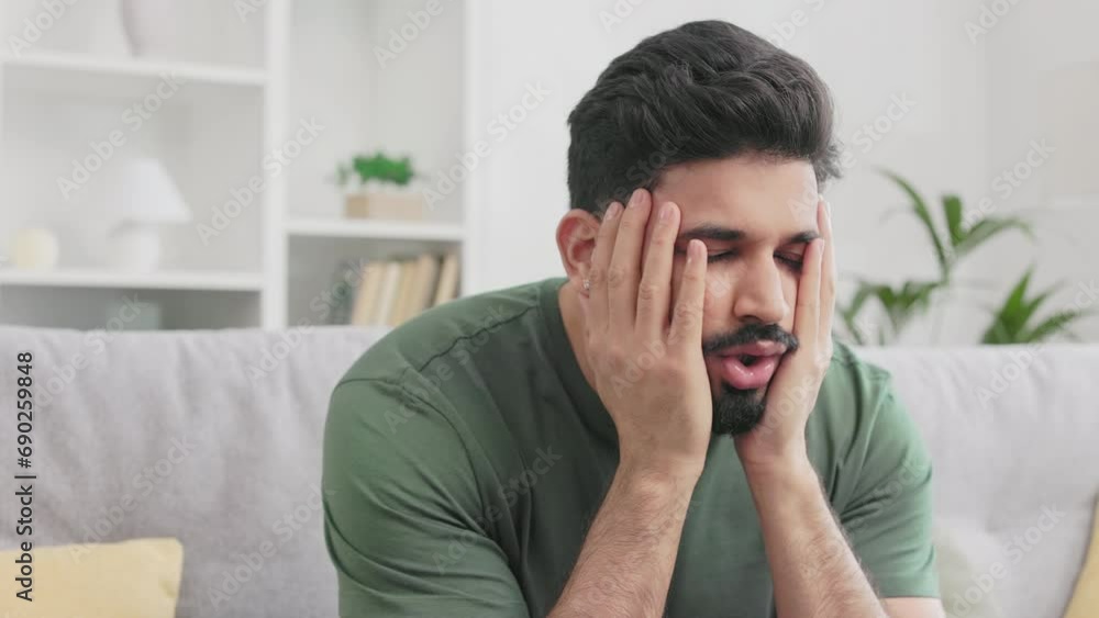 Stressed hindu guy touching head and gesturing from frustration while sitting on grey couch at living room. Bearded male person in casual wear experiencing negative emotions at home. Stressed hindu guy touching head and gesturing from frustration while sitting on grey couch at living room. Bearded male person in casual wear experiencing negative emotions at home.