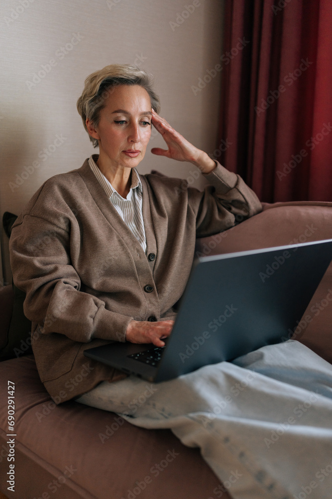 Vertical portrait of pensive Caucasian middle-aged business woman think business idea solution, working remotely, do freelance on-line job sitting on couch at home, looking at screen. Vertical portrait of pensive Caucasian middle-aged business woman think business idea solution, working remotely, do freelance on-line job sitting on couch at home, looking at screen.