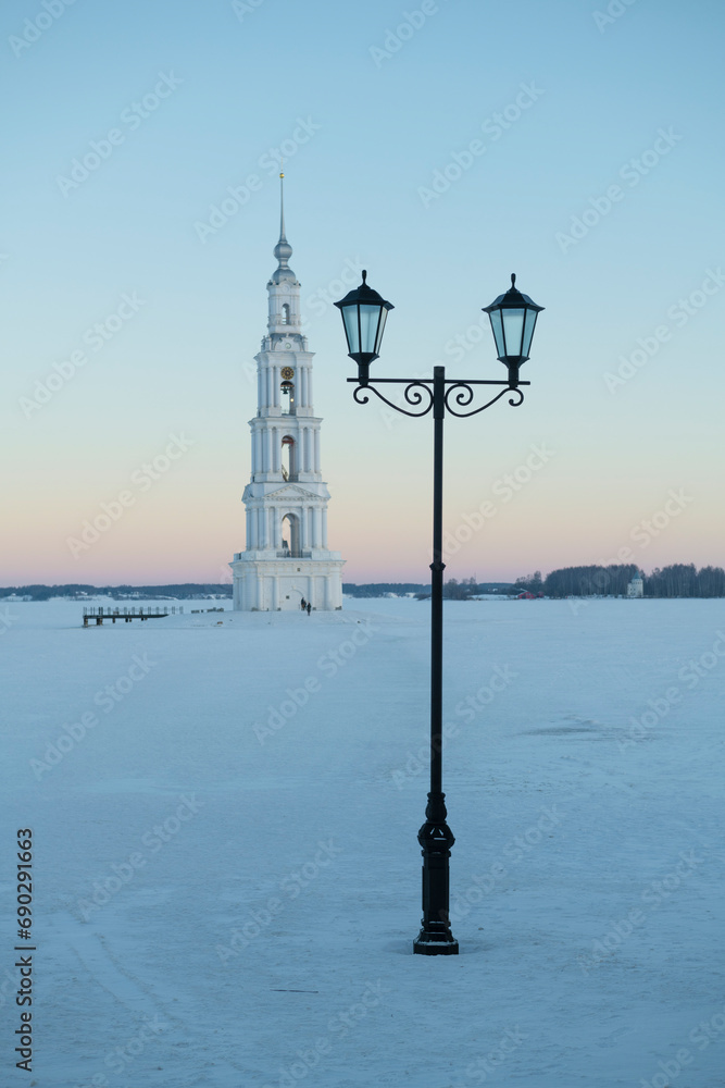 Fototapeta premium The city lantern against the background of an old bell tower on the frozen Uglich reservoir on January morning. Kalyazin. Tver regiont, Russia