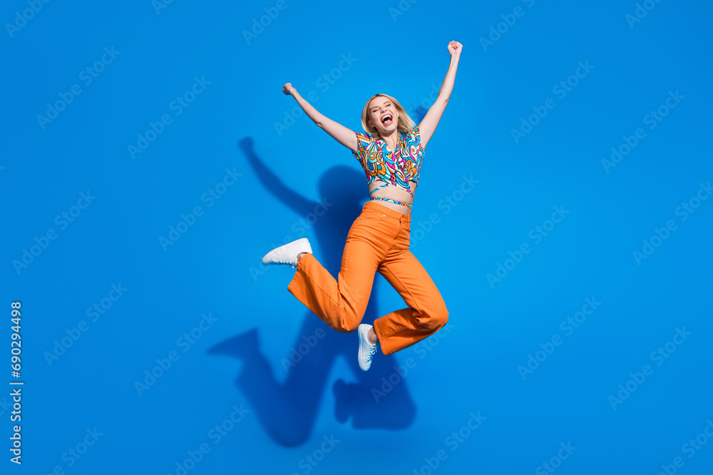 Full size photo of ecstatic optimistic girl wear blouse orange trousers jumping hands up win betting isolated on blue color background