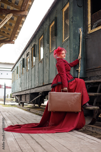 A beautiful girl in a burgundy suit of the last century and a hat with a veil stands with a suitcase in her hands near an old steam locomotive.Vintage portrait of the last century, retro journey