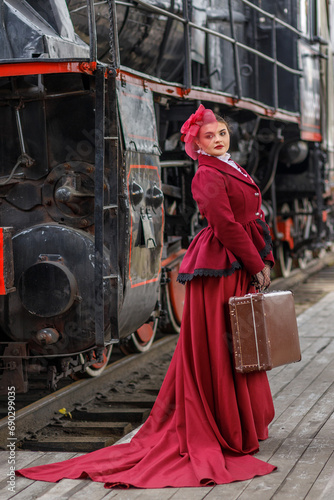 A beautiful girl in a burgundy suit of the last century and a hat with a veil stands with a suitcase in her hands near an old steam locomotive.Vintage portrait of the last century, retro journey