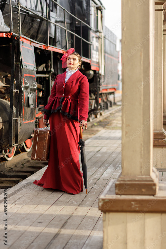 A beautiful girl in a burgundy suit of the last century and a hat with a veil stands with an umbrella and a cane near an old steam locomotive.Vintage portrait of the last century, retro journey