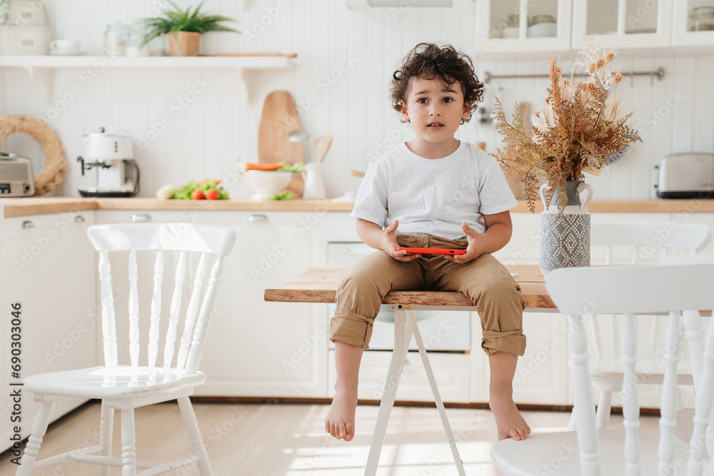 Handsome Brazilian little boy sitting on a kitchen table with phone, playing games at home. Curly caucasian kid using phone looking aside. Children and gadgets, childhood, Leisure at home, family. Handsome Brazilian little boy sitting on a kitchen table with phone, playing games at home. Curly caucasian kid using phone looking aside. Children and gadgets, childhood, Leisure at home, family.