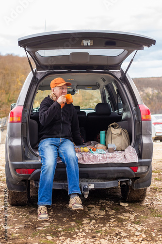 A Caucasian man sits in the trunk of a car and drinks coffee. Outdoor picnic. Road trip © Natalia