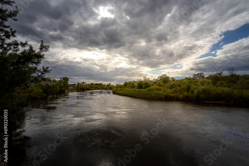 The sky with dramatic clouds is reflected in the river. A wide, full-flowing river in a bend. Pine branches in the foreground