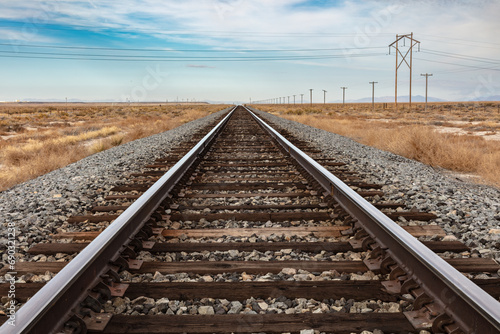 Closeup of railroad tracks in central Utah. Stretching straight to the horizon. Telephone poles to the right. Blue cloudy sky beyond.
