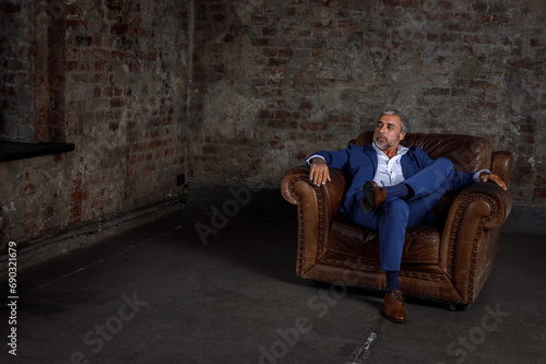 A handsome middle-aged man in a blue formal suit poses sitting on a leather armchair. Portrait of a businessman. A handsome middle-aged man poses on a brick background with a place to copy