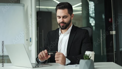 Smiling young caucasian male holding credit card and entering data into modern laptop in designer office. Competent bearded businessman doing shopping online and making payment purchase indoors. photo