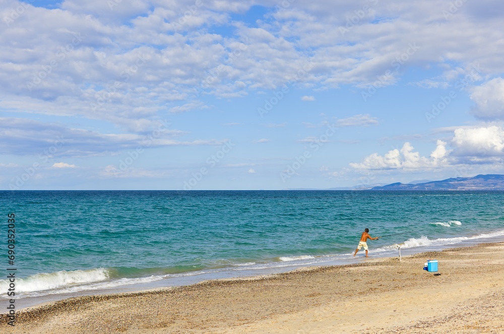 A Sardinian fisherman with a rod on Platamona Beach, Sardinia, Italy. The photo captures the beach, distant mountains, and beautiful clouds against the blue sky.