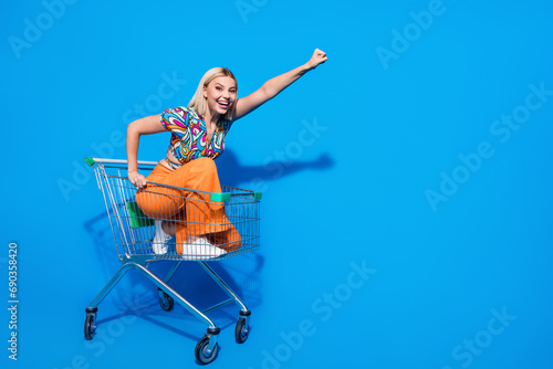 Full size photo of cheerful girl wear blouse sitting in trolley raisng fist up riding at supermarket isolated on blue color background