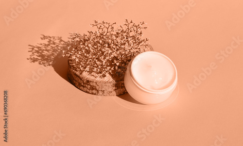 Peach fuzz is color of year 2024, tinted image. Wooden podium and dry flower with a white jar of cosmetic products.