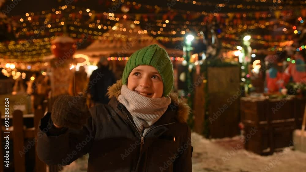 happy boy in winter clothes holding a burning sparkler, having a joyful time at the Christmas market in the evening in the city center happy boy in winter clothes holding a burning sparkler, having a joyful time at the Christmas market in the evening in the city center