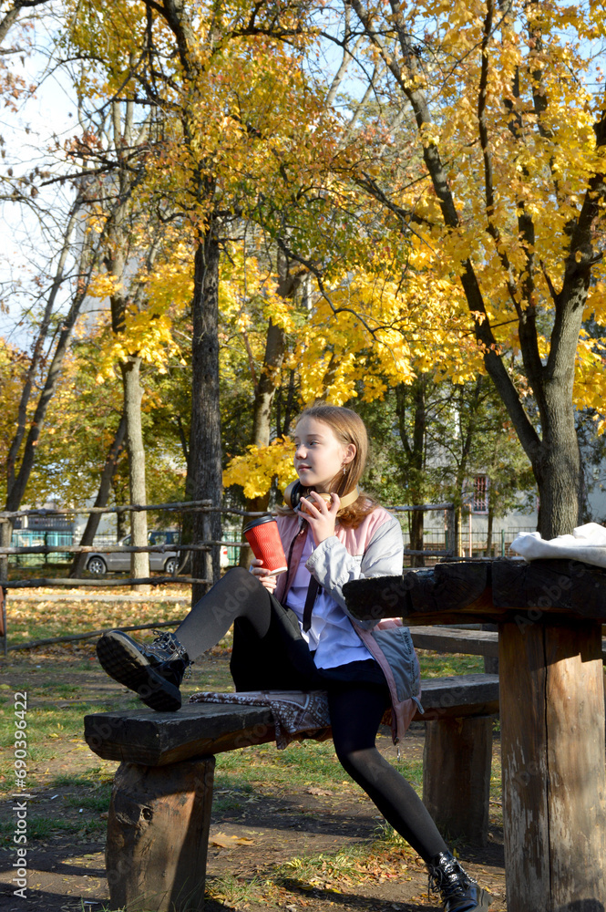a girl sits at a table in an autumn park, headphones are on her head, and a cup of a warming drink is nearby. child listens to music or audio book. hobbies, recreation and entertainment a girl sits at a table in an autumn park, headphones are on her head, and a cup of a warming drink is nearby. child listens to music or audio book. hobbies, recreation and entertainment