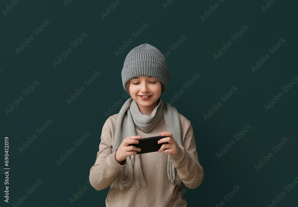 Cute little happy boy in winter clothes playing with phone on dark green background Cute little happy boy in winter clothes playing with phone on dark green background