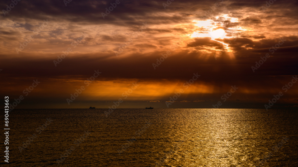 Fototapeta premium Divine dramatic seascape with dark clouds, a glowing sun over the horizon, and ships in the distance