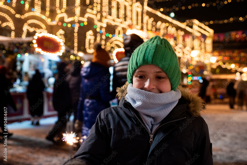 happy boy in winter clothes holding a burning sparkler, having a joyful time at the Christmas market in the evening in the city center