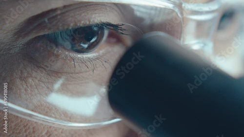 Brown eyes of female scientist in safety glasses looking through microscope oculars in laboratory photo