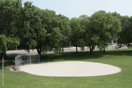 gray gravel circle baseball diamond with road behind and trees, silver diamond fence at home base plate surrounded by grass, shot from high vantage point looking down photo