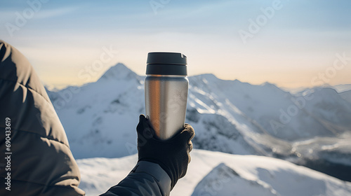 Hand in glove holding a thermos with a majestic mountain range at sunrise.