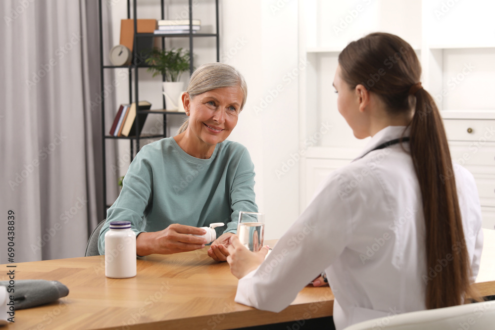Young healthcare worker giving glass of water to senior woman with pills at wooden table indoors Young healthcare worker giving glass of water to senior woman with pills at wooden table indoors