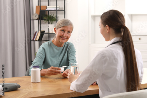 Young healthcare worker giving glass of water to senior woman with pills at wooden table indoors