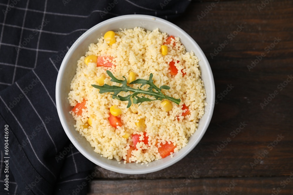 Tasty couscous with pepper, corn and arugula in bowl on wooden table, top view