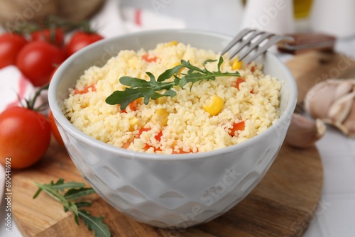 Bowl of tasty couscous with pepper, corn, arugula and fork on white table, closeup