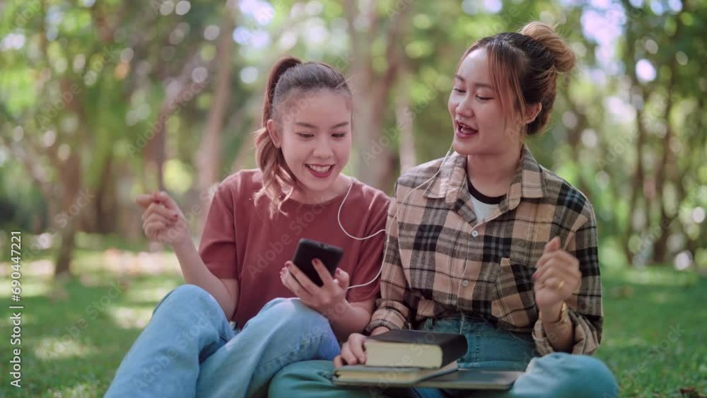 Asian female college student is using her smartphone to listen to music through headphones and sharing the experience with her friend, aiming to unwind and relax in a garden with lush green trees. Asian female college student is using her smartphone to listen to music through headphones and sharing the experience with her friend, aiming to unwind and relax in a garden with lush green trees.