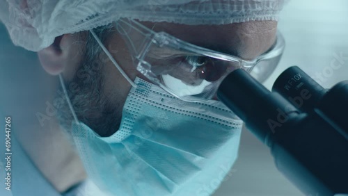 Close up view of focused chemist wearing face mask, safety glasses and medical hat looking through microscope and writing down results of lab research
