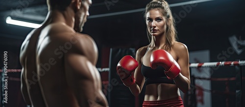 Young woman trains in boxing ring with partner near red punching bag and other equipment.