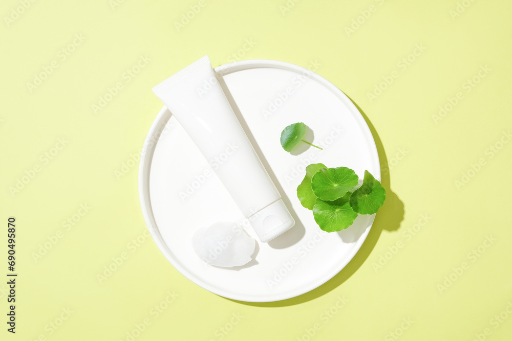 An unlabeled cosmetic tube and fresh pennywort leaves are displayed on a white ceramic plate on a pastel background. Vegan cosmetic concept with pennywort extract.