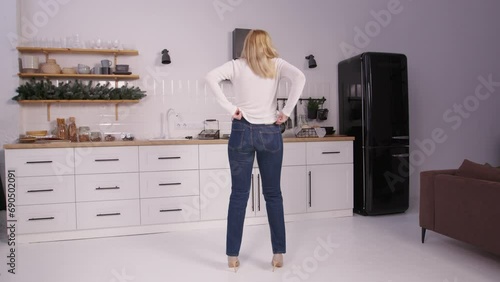 Rear view of disappointed pretty middle aged woman in high heels trying to squeeze into old tight jeans after gaining weight, expressing frustration and despair while standing in domestic kitchen. photo