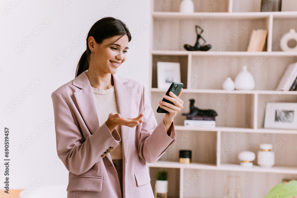 Cheerful Asian brunette business woman in pink suit holds phone makes video call smiles at home against bookshelf. Happy Korean girl surprised by great news reads message. Home office. Cheerful Asian brunette business woman in pink suit holds phone makes video call smiles at home against bookshelf. Happy Korean girl surprised by great news reads message. Home office.