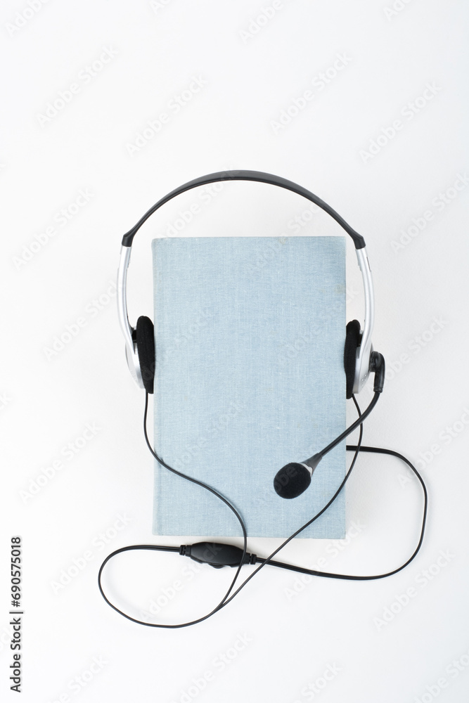 Audiobook on white background. Headphones put over blue hardback book, empty cover, copy space for ad text. Distance education, e-learning concept. Top view Audiobook on white background. Headphones put over blue hardback book, empty cover, copy space for ad text. Distance education, e-learning concept. Top view
